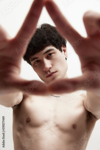 Through the lens of perception. Portrait of handsome young guy posing shirtless against white studio background. Looking through hands. Concept of male body aesthetics, style, health, men's beauty