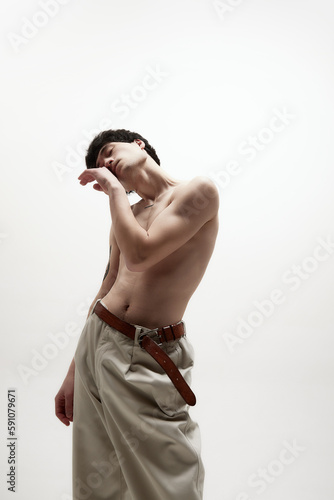 Self-care. Portrait of young handsome man with fit body  posing shirtless in beige pants against white studio background. Concept of male body aesthetics  style  fashion  health  men s beauty