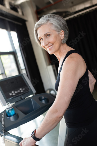 happy senior woman with grey hair running on treadmill next to sports bottle with water.