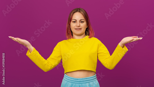 Attractive smiling woman with pink hair looking at camera and showing balance with hands, wear a yellow t-shirt, stand pink background