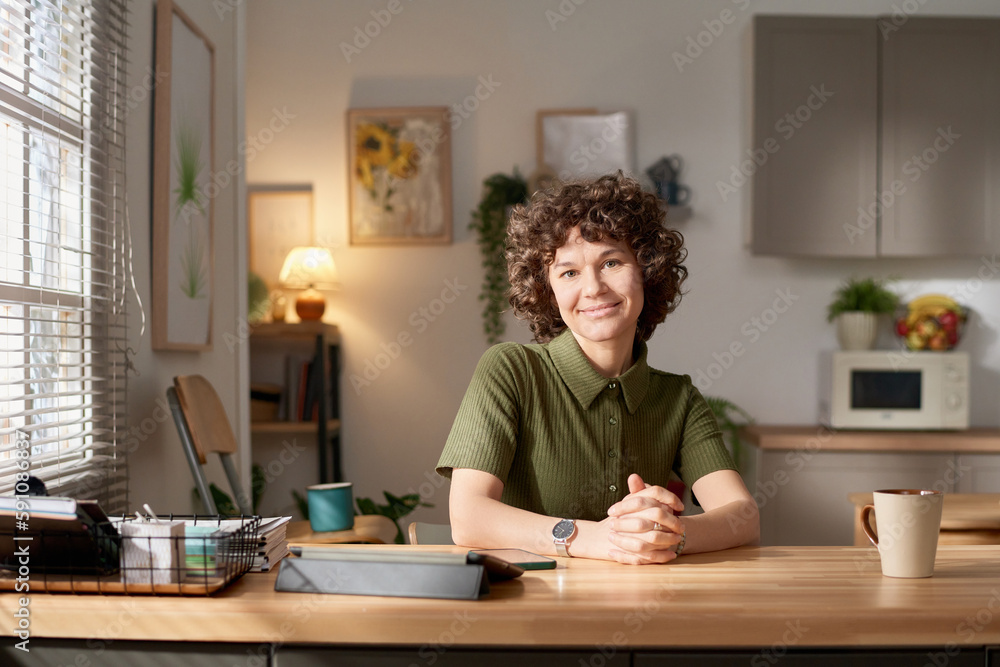 Portrait of young woman with curly hair looking at camera while sitting at kitchen table with tablet pc and cup of coffee Portrait of young woman with curly hair looking at camera while sitting at kitchen table with tablet pc and cup of coffee