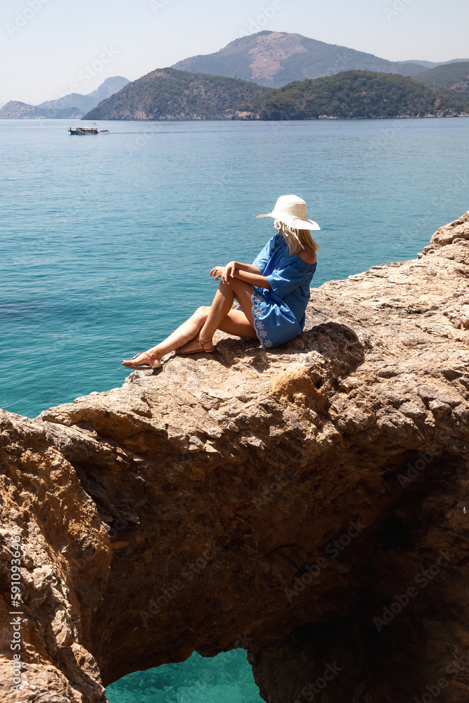 Young pretty woman in blue beachwear and hat sitting on rock at tropical azure ocean background, summer vacation. Lovely tourist lady enjoy rest on sea. Travel holidays concept. Copy ad text space