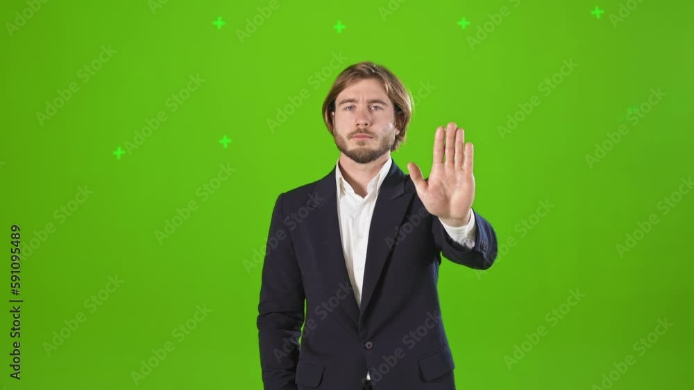 Front view of handsome, young man standing in studio, raising hand, showing stop. Male wearing costume, shaking head, denying, objecting. Isolated on green studio background. Front view of handsome, young man standing in studio, raising hand, showing stop. Male wearing costume, shaking head, denying, objecting. Isolated on green studio background.