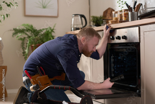 Professional worker examining the problem with oven and repairing it during his visit to home