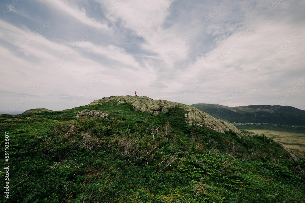 Distant shot of a traveler standing on the peak of a hill with green plants growing on it