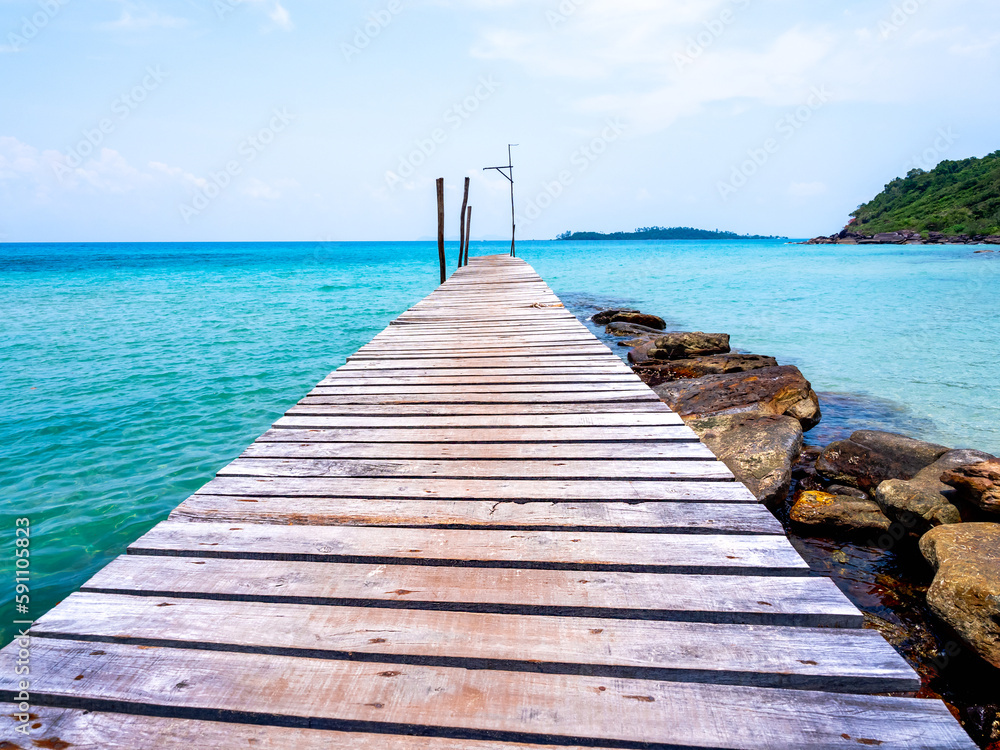 Obraz premium Wooden bridge heading to the blue sea. Brown wood plank pathway bridge on the beach sand with rocks at the local port in the island on sunny day. Seascape summer holiday background.