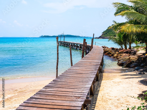 Wooden bridge heading to the blue sea. Brown wood plank pathway bridge on the beach sand with rocks and coconut palm tree at the local port in island on sunny day. Seascape summer holiday background. © tete_escape