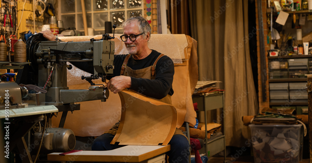 Naklejka premium senior tanner sitting sewing leather back panel with a sewing machine for a chair made by him in workshop