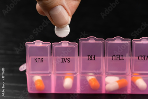 Obraz na plátne Close-up of a woman with her hand taking pills from a pink pillbox or sorting pills and vitamins for daily medication on a black table