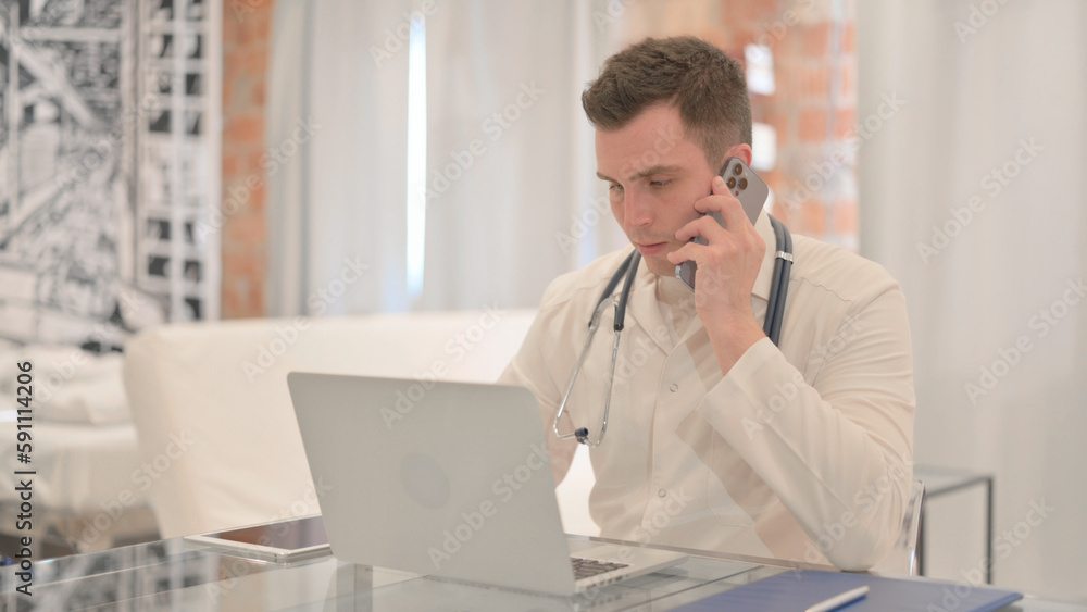 Male Doctor Talking on Phone while Working on Laptop in Clinic