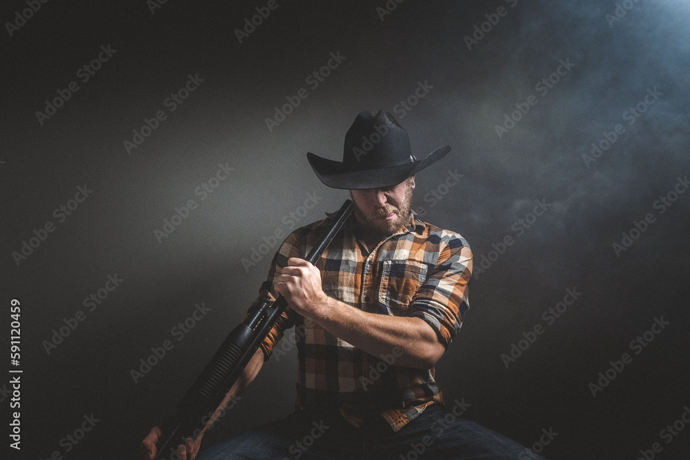 Rugged man with beard and a shotgun in cowboy hat. Plaid shirt. Blue jeans. Studio session. Rugged man with beard and a shotgun in cowboy hat. Plaid shirt. Blue jeans. Studio session.