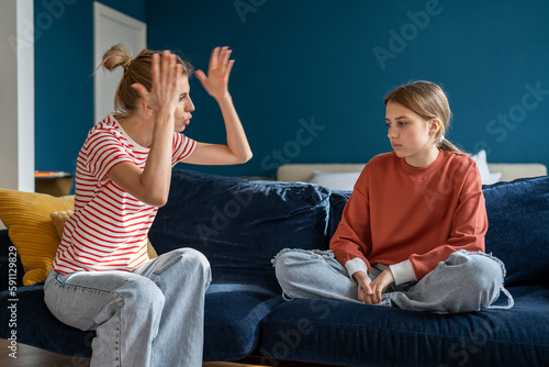 Young angry emotional mother screaming at troubled teen daughter, furious parent mom dealing with teenage behavior problems, yelling at upset adolescent girl while sitting together on sofa at home