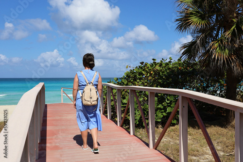 Picturesque view to path to tropical beach with palm trees. Lonely woman in blue dress walking towards ocean coast, tourist resort on Caribbean island