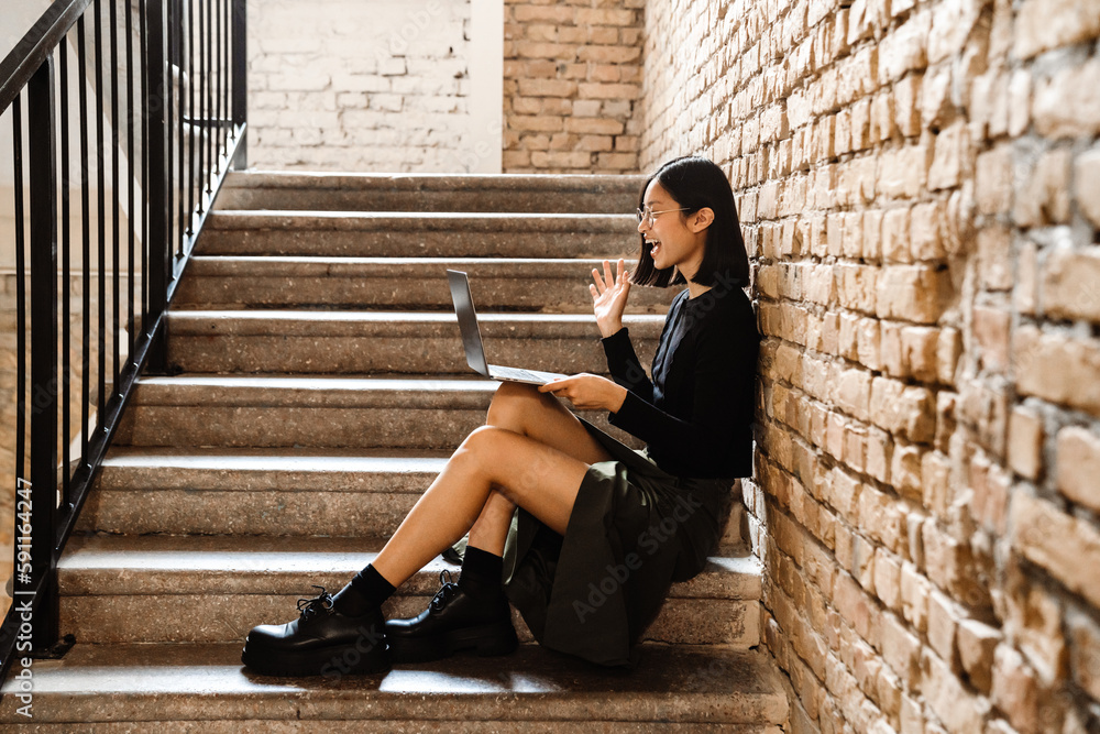 Cheerful asian woman making video call via laptop while sitting on stairs indoors Cheerful asian woman making video call via laptop while sitting on stairs indoors