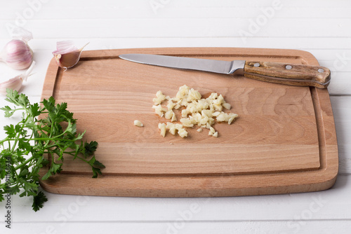 Wooden board with chopped garlic, a whole clove of garlic, parsley and a knife on a white background, top view. Cooking homemade vegetable stew or other dish