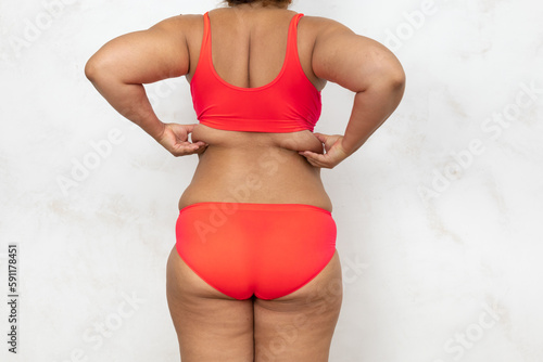 Overweight woman standing with back pinch fat folds by hands, free copy space, white background. Bare woman in red underwear show large cellulite body. Plus size people, weight loss, self acceptance.