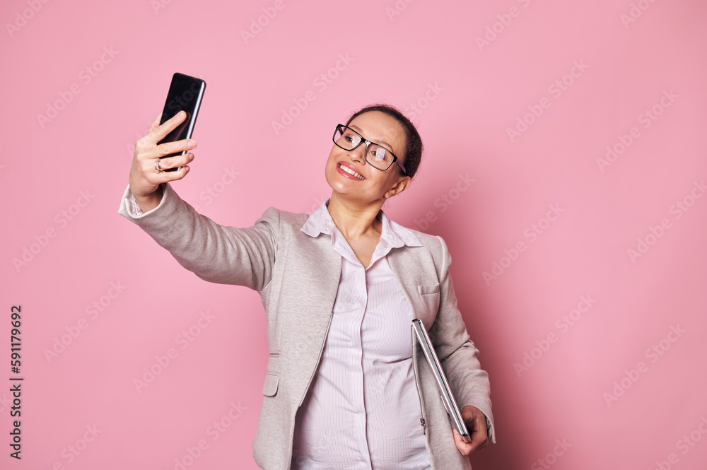 Attractive middle-aged multi-ethnic pregnant woman, business person wearing casual clothes and trendy glasses, carrying laptop and taking selfie on her smartphone on isolated pink background. Ad space Attractive middle-aged multi-ethnic pregnant woman, business person wearing casual clothes and trendy glasses, carrying laptop and taking selfie on her smartphone on isolated pink background. Ad space