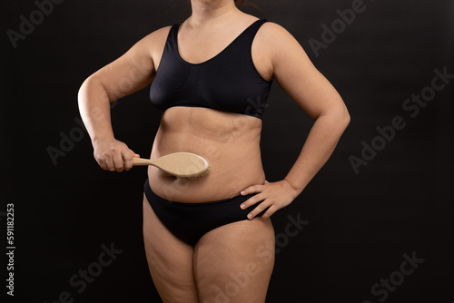 Plus size woman in black underwear massaging belly with brush. Flaunt figure imperfections. Dry anti cellulite massage. Studio portrait over black background. Concept of skin care, body positive.