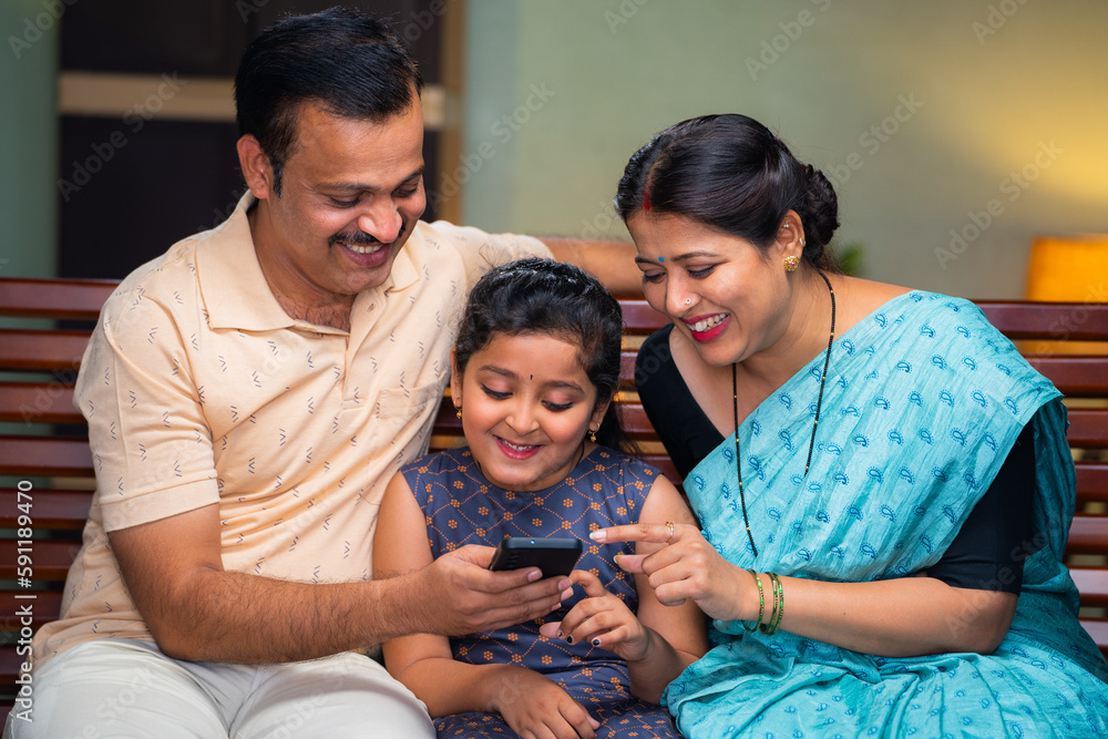 Happy smiling couple with daughter watching mobile phone while sitting on sofa at home - concept of family time, cyberspace and caring parents