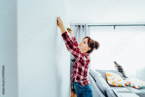 Young woman in plaid shirt doing measuring with a measure tape on the wall. Girl wants to put a picture on the wall at home. Housekeeping work. Doing repair herself. DIY, equality in work concept. photo