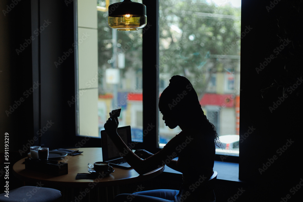 Backlit silhouette of young busy African-American woman with long dark braids wearing roll-neck sweater, sitting at table with cup of coffee in cafe, working typing on laptop, giving credit card. 