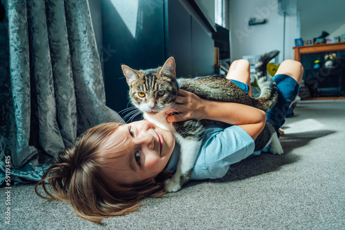 Portrait of a Happy smiling kid at home playing with a kitty cat. Caucasian boy lying on a carpeted floor holding his cat in the living room. Life with a pet. Best friends.Selective focus.