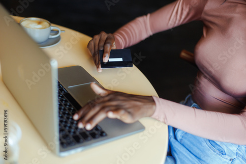 Cropped photo of African-American woman wearing pink sweater, sitting at table with cup of coffee in cafe, working, doing online shopping on laptop, holding credit card. E-commerce, internet banking.