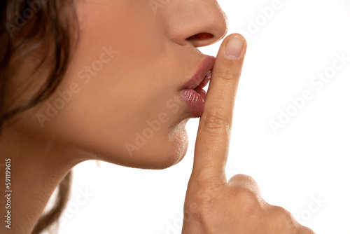 Cropped image of a young woman holding index finger at her lips, saying 'shh' on a white background. side profile view