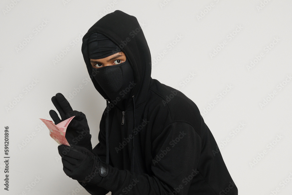 Portrait of mysterious man wearing black hoodie and mask stealing one hundred thousand rupiah successfully from victim. Isolated image on gray background Portrait of mysterious man wearing black hoodie and mask stealing one hundred thousand rupiah successfully from victim. Isolated image on gray background