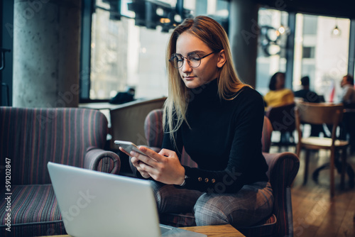 Focused female freelancer using smartphone while sitting in cafe with laptop