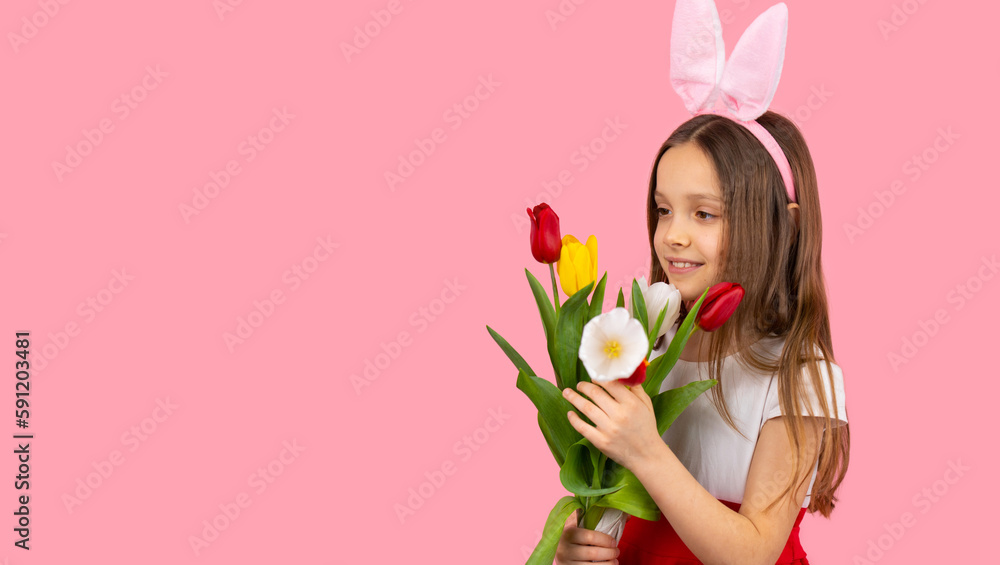 Little blond pretty girl wearing bunny ears on head in red-white dress looking at the flowers she holding in her hands on pink background and copy space. Little blond pretty girl wearing bunny ears on head in red-white dress looking at the flowers she holding in her hands on pink background and copy space.