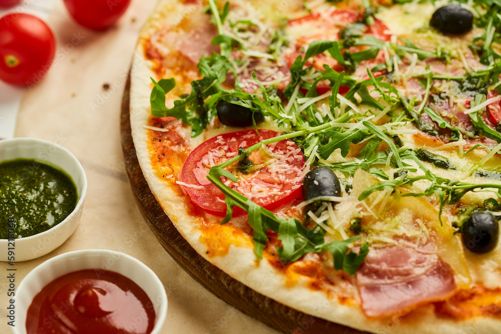 Freshly baked pizza with ham, rukkola, sauce pesto and olives served on wooden background with tomatoes, sauces and herbs. Food delivery concept. Restaurant menu Freshly baked pizza with ham, rukkola, sauce pesto and olives served on wooden background with tomatoes, sauces and herbs. Food delivery concept. Restaurant menu