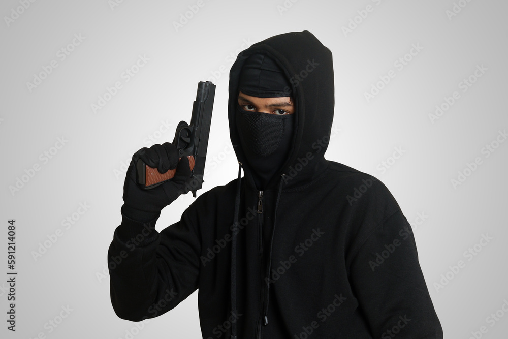 Mysterious man wearing black hoodie and mask holding a pistol, shooting with a gun. Isolated image on gray background Mysterious man wearing black hoodie and mask holding a pistol, shooting with a gun. Isolated image on gray background