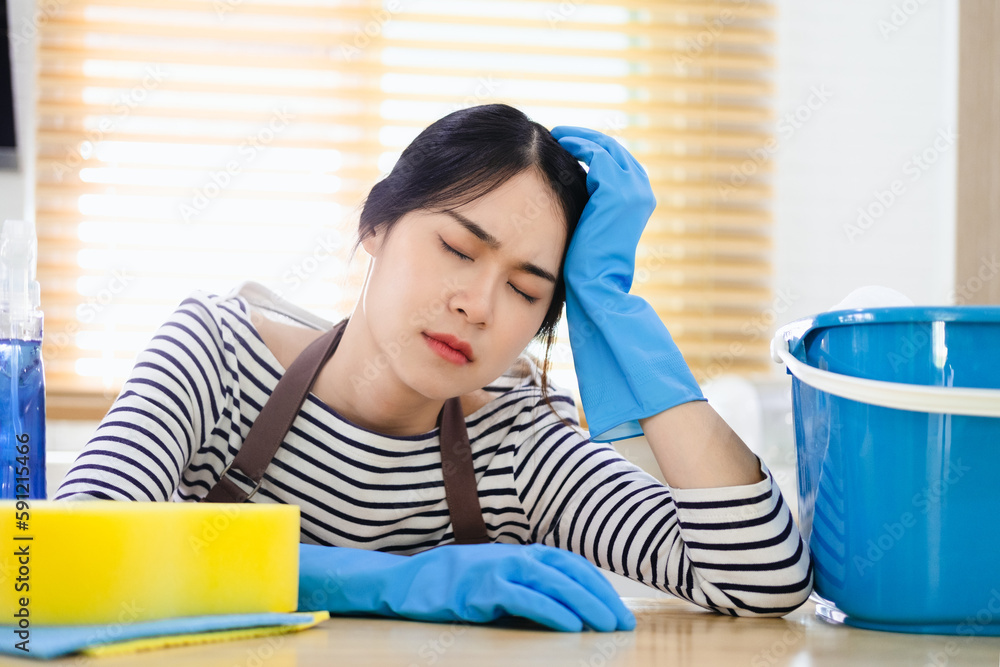 Beautiful Asian housewife feeling tired after doing housework. Exhausted housekeeper in brown apron and blue rubber gloves sitting in kitchen with home cleaning products. Beautiful Asian housewife feeling tired after doing housework. Exhausted housekeeper in brown apron and blue rubber gloves sitting in kitchen with home cleaning products.