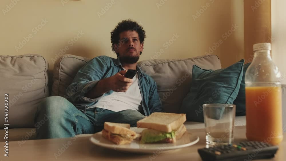 Young man sits in the living room with remote control in his hands, eating lunch Young man sits in the living room with remote control in his hands, eating lunch