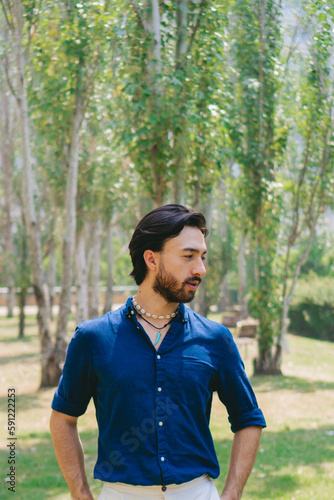 Well dressed bearded young Latin man standing in a city park looking to the side.