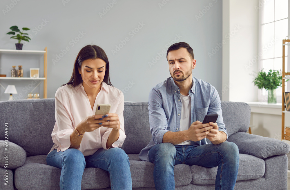 Jealous man spying looking at his girlfriend phone. Couple sitting on couch with smartphones, suspicious husband trying to read messages at wife smartphone while she texting message and smiling - obrazy, fototapety, plakaty 