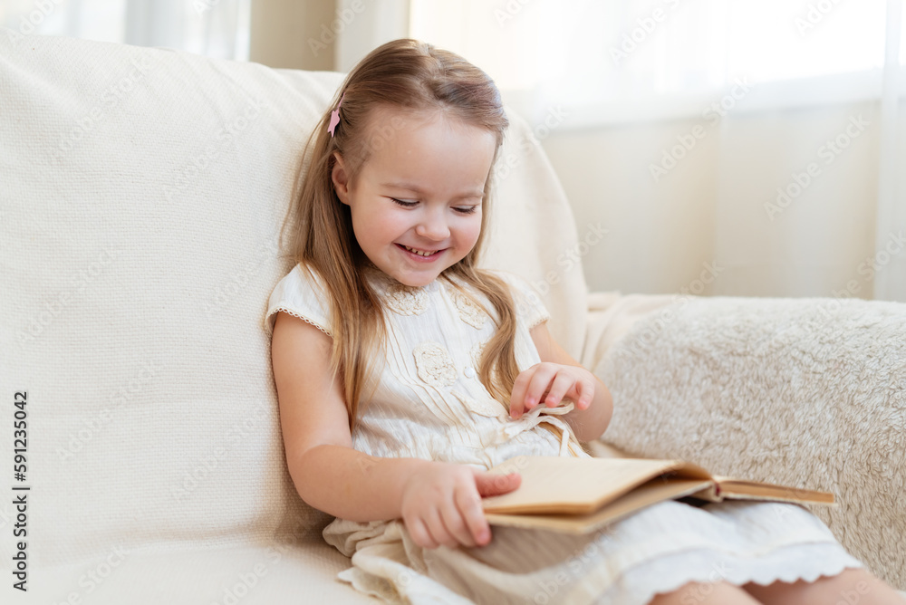Little cute blond girl reading book siting on a sofa. Child reading, dreaming and imagination development. International Children's Book Day. Kids Love to reading. World Book Day Little cute blond girl reading book siting on a sofa. Child reading, dreaming and imagination development. International Children's Book Day. Kids Love to reading. World Book Day