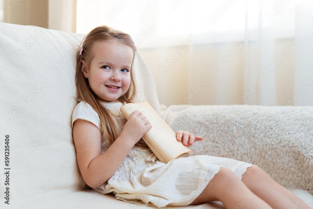 Little cute blond girl reading book siting on a sofa. Child reading, dreaming and imagination development. International Children's Book Day. Kids Love to reading. World Book Day Little cute blond girl reading book siting on a sofa. Child reading, dreaming and imagination development. International Children's Book Day. Kids Love to reading. World Book Day