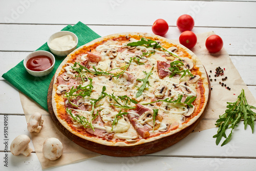 Freshly baked pizza with ham, rukkola and mushrooms served on wooden background with tomatoes, sauces and herbs. Food delivery concept. Restaurant menu