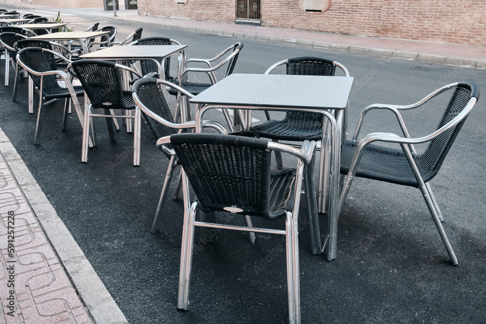 Black and white metal tables and chairs near a street cafe.Rows of empty aluminium tables and chairs cleaned and ready for the opening time of a cafe. Black and white metal tables and chairs near a street cafe.Rows of empty aluminium tables and chairs cleaned and ready for the opening time of a cafe.