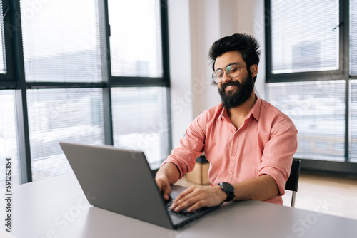 Cheerful Indian businessman in eyeglasses working on laptop sitting at desk in light coworking office room on background of window. Bearded business man in casual clothes looking at computer screen.