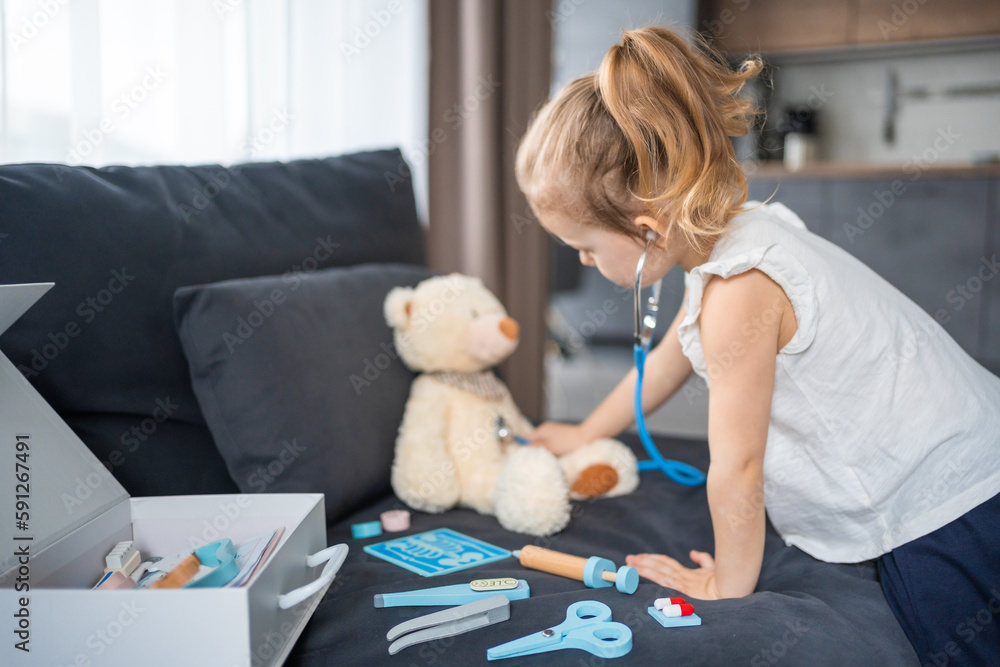 Little girl playing doctor with toys and teddy bear on the sofa in living room at home Little girl playing doctor with toys and teddy bear on the sofa in living room at home