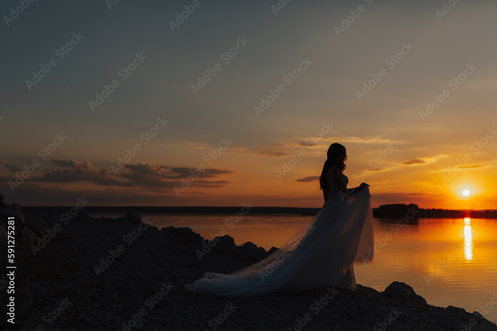Silhouette of young bride in the long dress  walking along the sea promenade. Summer, sunset time. Outdoor wedding ceremony on the sea.