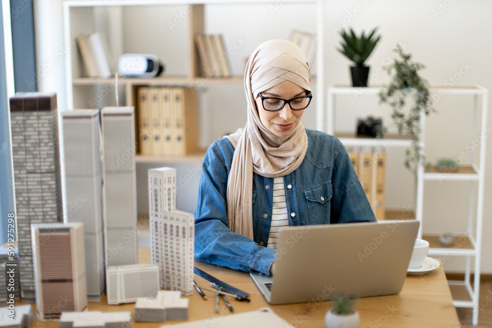 Focused bespectacled muslim female in hijab typing on portable computer at office desk with building models on it. Experienced architect developing urban design using modern gadgets and drawing tools. Focused bespectacled muslim female in hijab typing on portable computer at office desk with building models on it. Experienced architect developing urban design using modern gadgets and drawing tools.
