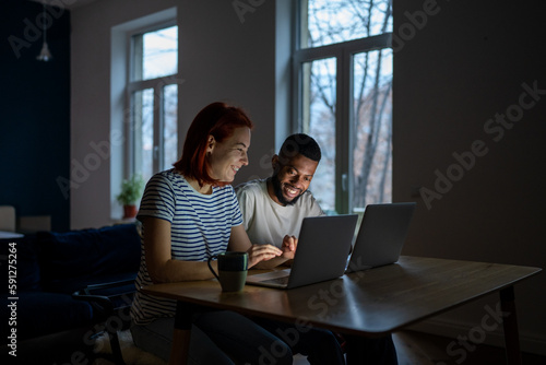 Modern smiling multiracial family couple wife and husband enjoy freelancing, sit together at table working on laptops, happy African man and Caucasian woman run successful online business from home