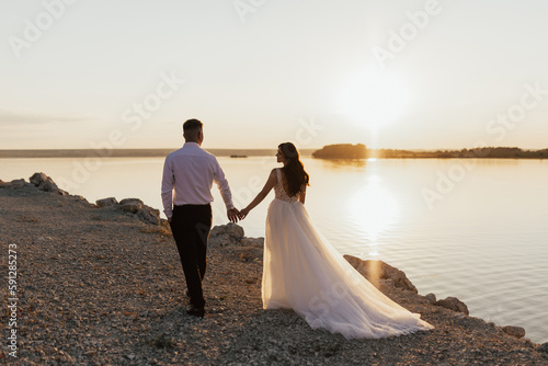 Couple holding hands, looking each other and walking on the beach at sunset. Lovely couple enjoying sunset at the beach. Wedding ceremony at sea.