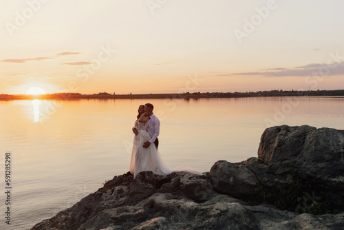 Newlyweds hugging on the rocky shore of the sea overlooking the sunset. Outdoor wedding ceremony on the sea.