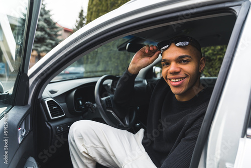 Young man sitting in a driver seat with an open car door and smiling to the camera. High quality photo © PoppyPix