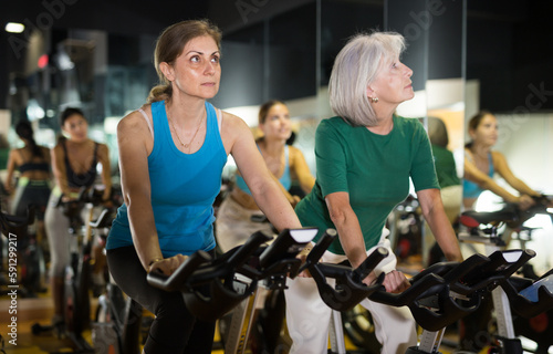 Two confident senior women warming up on bikes in spin class at fitness studio